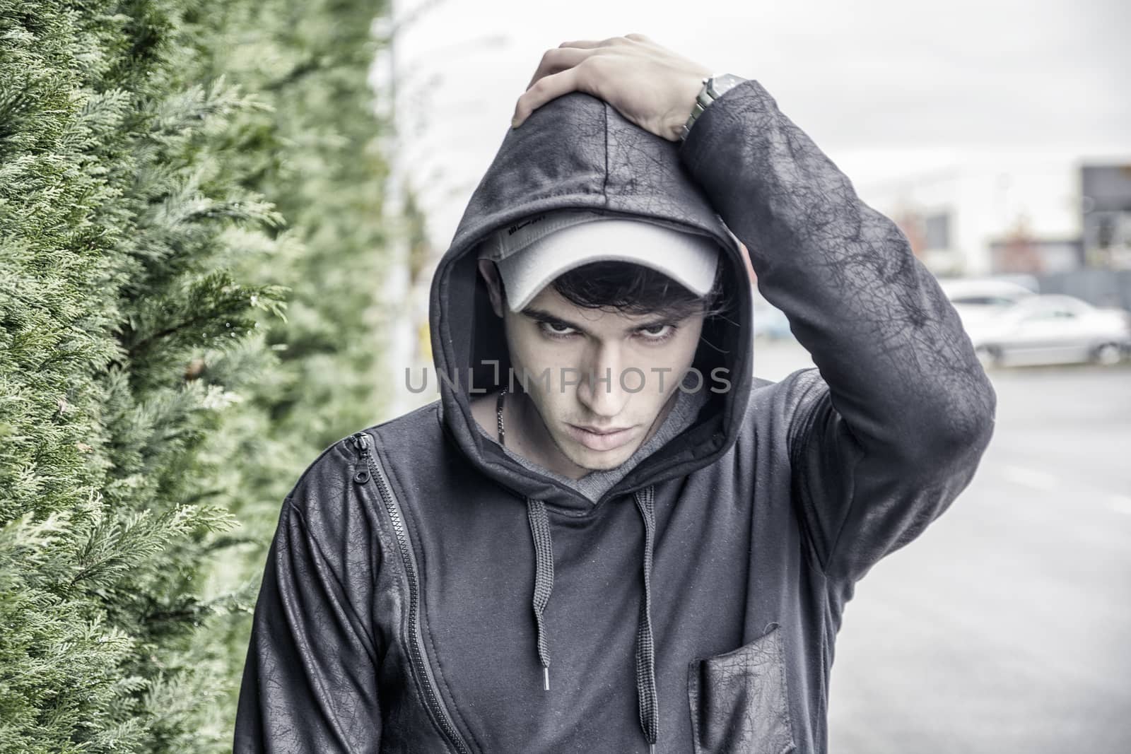 Handsome young man in black hoodie sweater standing outdoor in street looking at camera, hand over hood