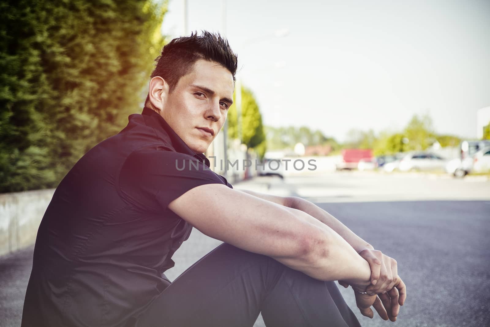 Handsome muscular young man in black shirt sitting outdoor in street looking at camera