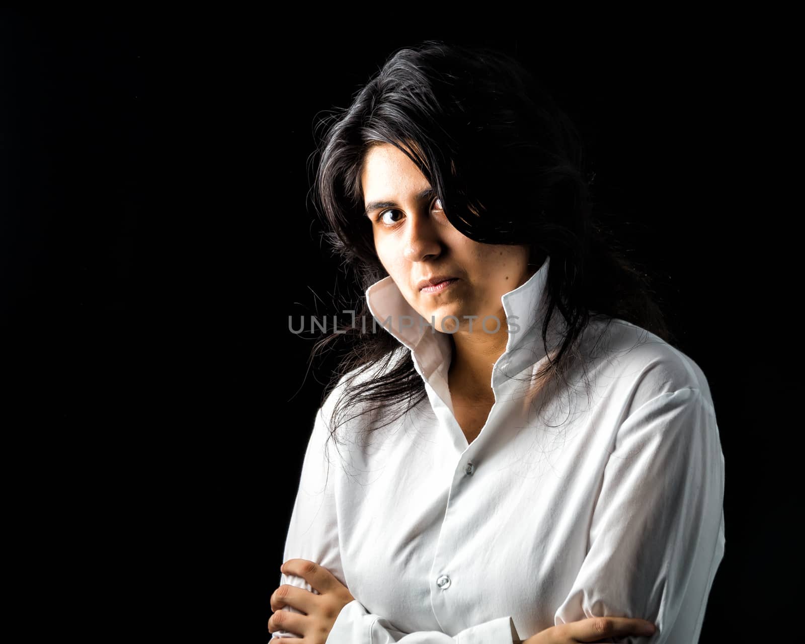 Latina teen in white long sleeved shirt standing in front of black backdrop with arms crossed and looking at the camera