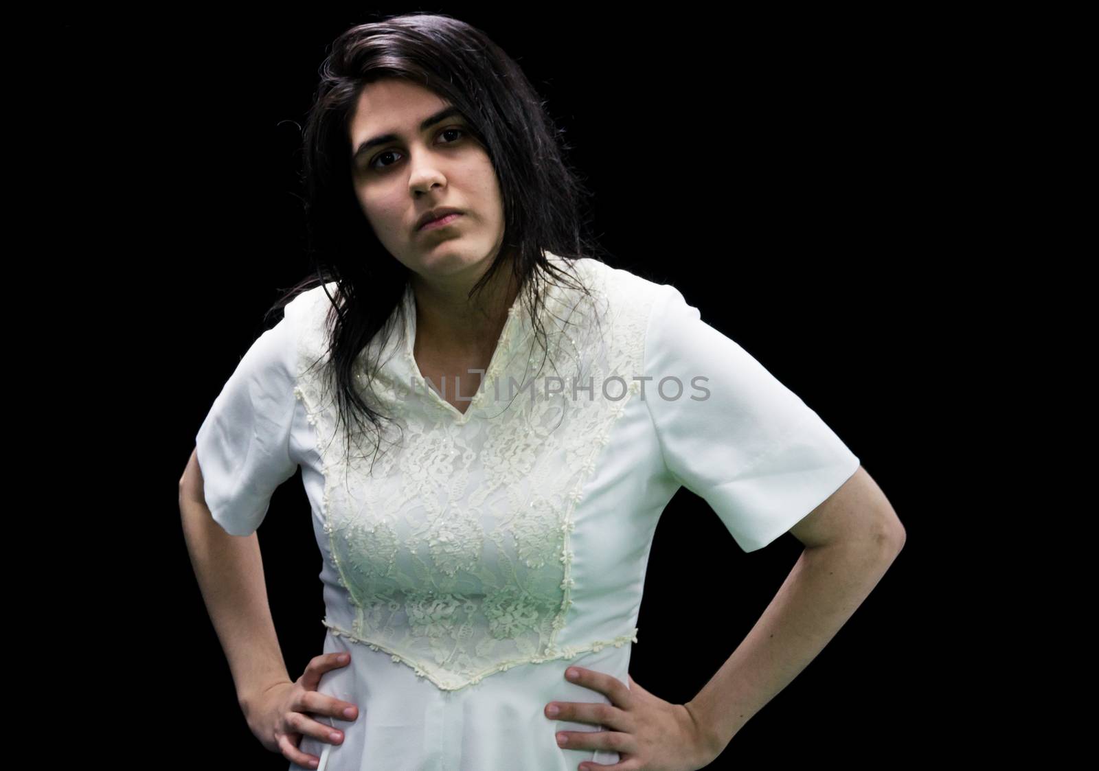 Latina teenager in white standing in front of a black background with hands on hips and a serious look on her face