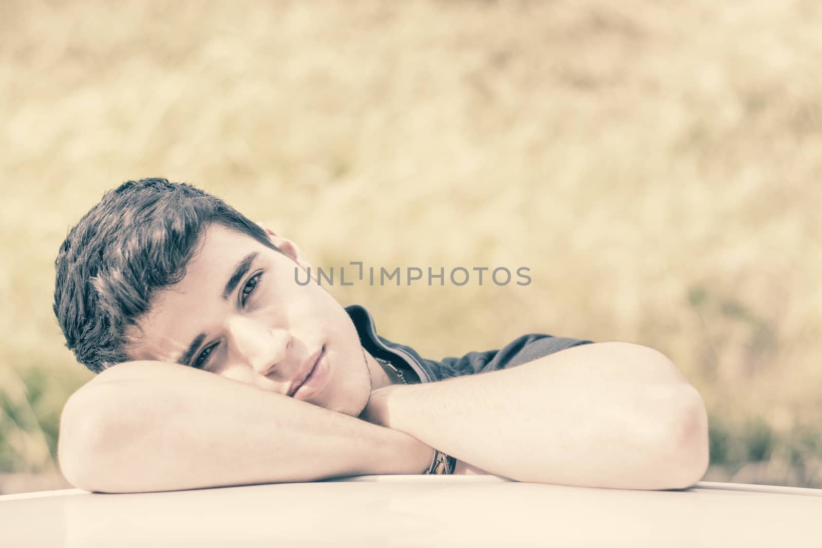 Head and arms shot of handsome attractive young man looking at camera outdoor, leaning on flat surface with head resting on hands