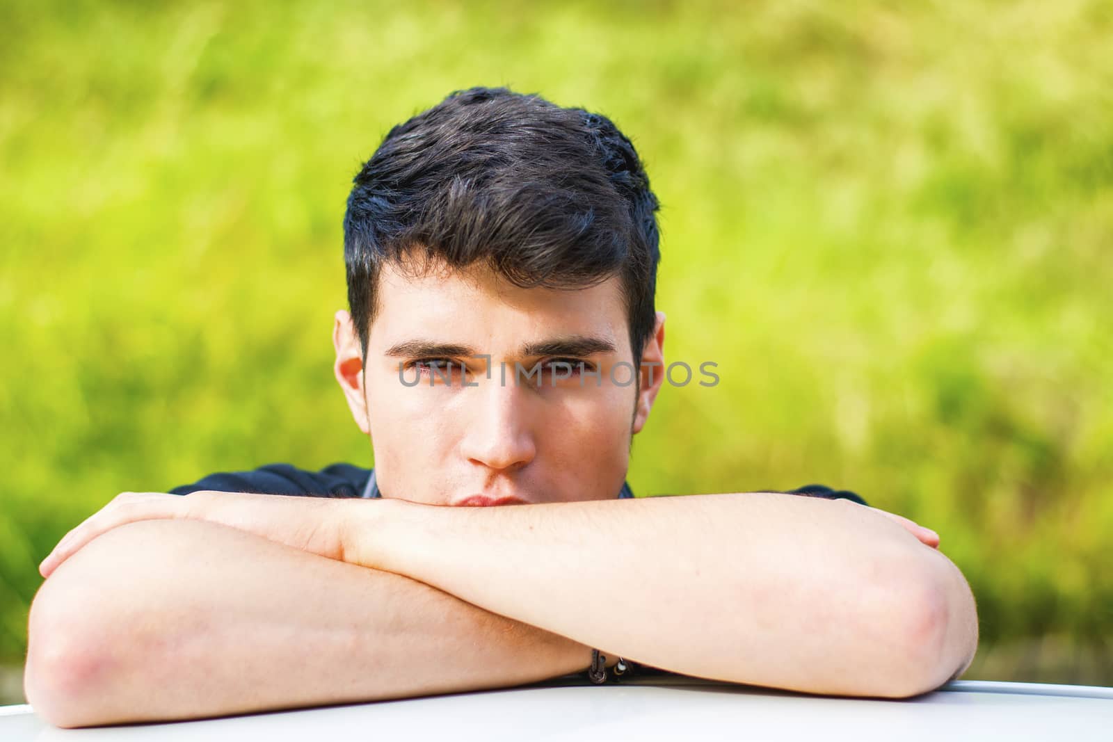 Head and arms shot of handsome attractive young man looking at camera outdoor, leaning on flat surface with head resting on hands