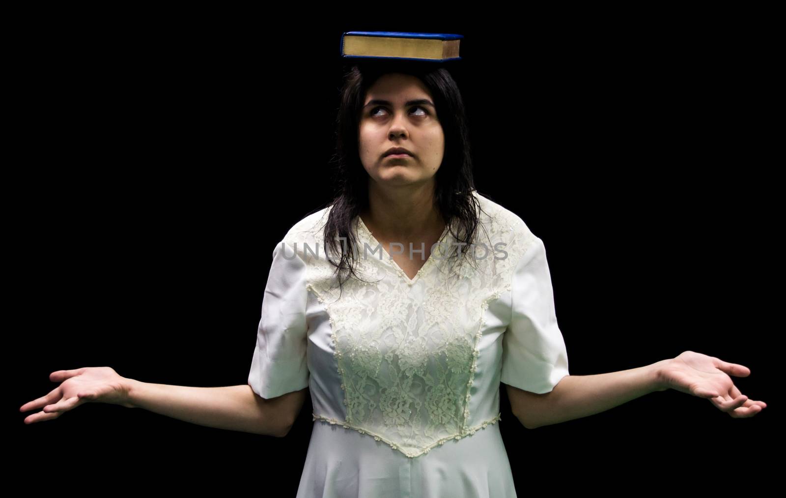 Latina teenager in white dress standing in front of black background balancing book on her head and rolling her eyes