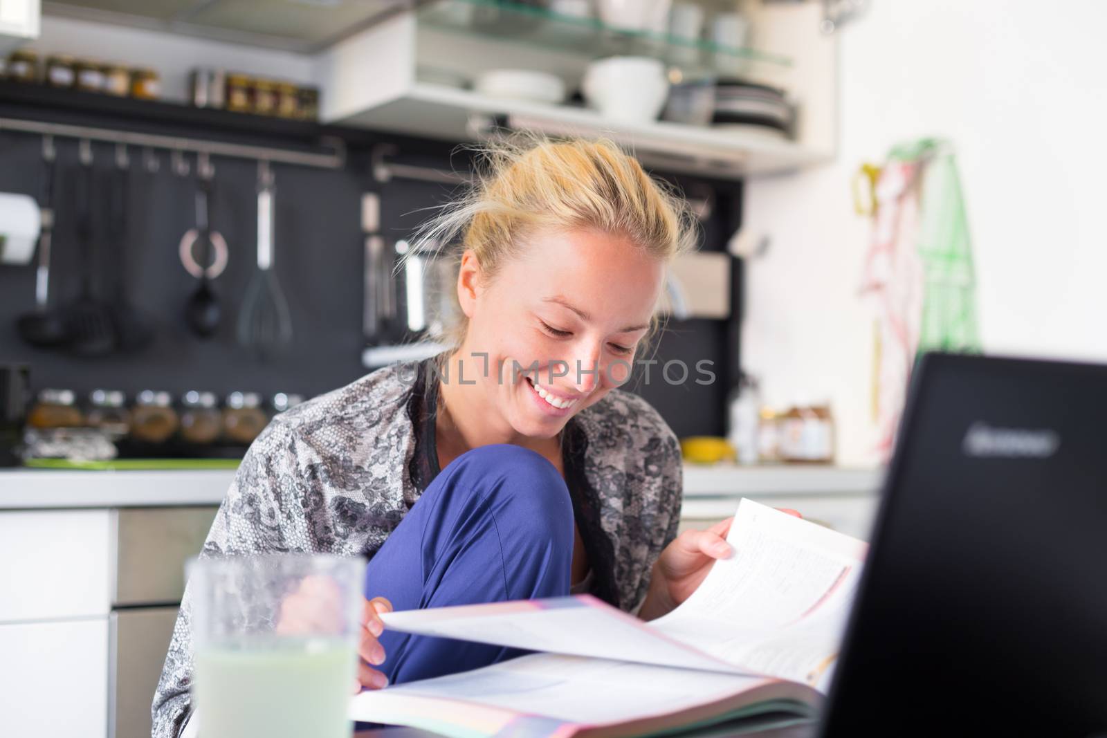 Female student in her casual home clothing working and studying remotly from her small flat in the morning. Home kitchen in the background. Great flexibility of web-based courses and study programmes.