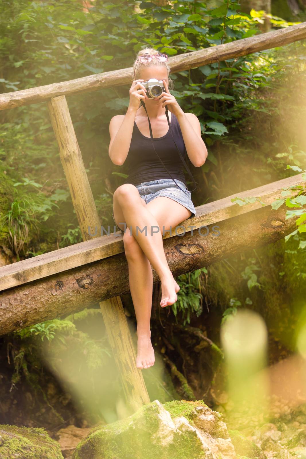 Beautiful barefooted blonde caucasian girl wearing jeans shorts an sporty black sleeveless t-shirt, sitting on a vintage wooden bridge, taking photos with retro camera.