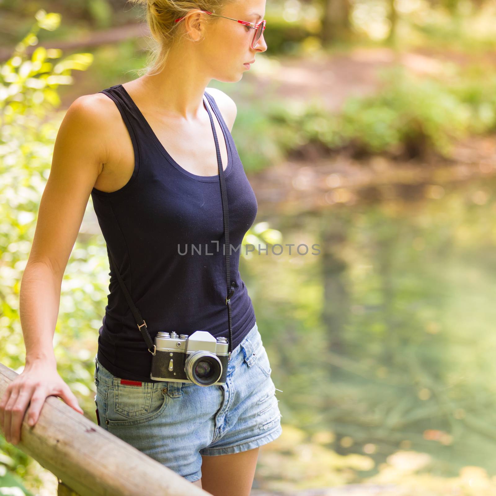 Beautiful blonde caucasian girl wearing jeans shorts an sporty black sleeveless t-shirt, outdoors in nature, carrying vintage camera over her shoulder. Healthy active lifestyle. Square composition.