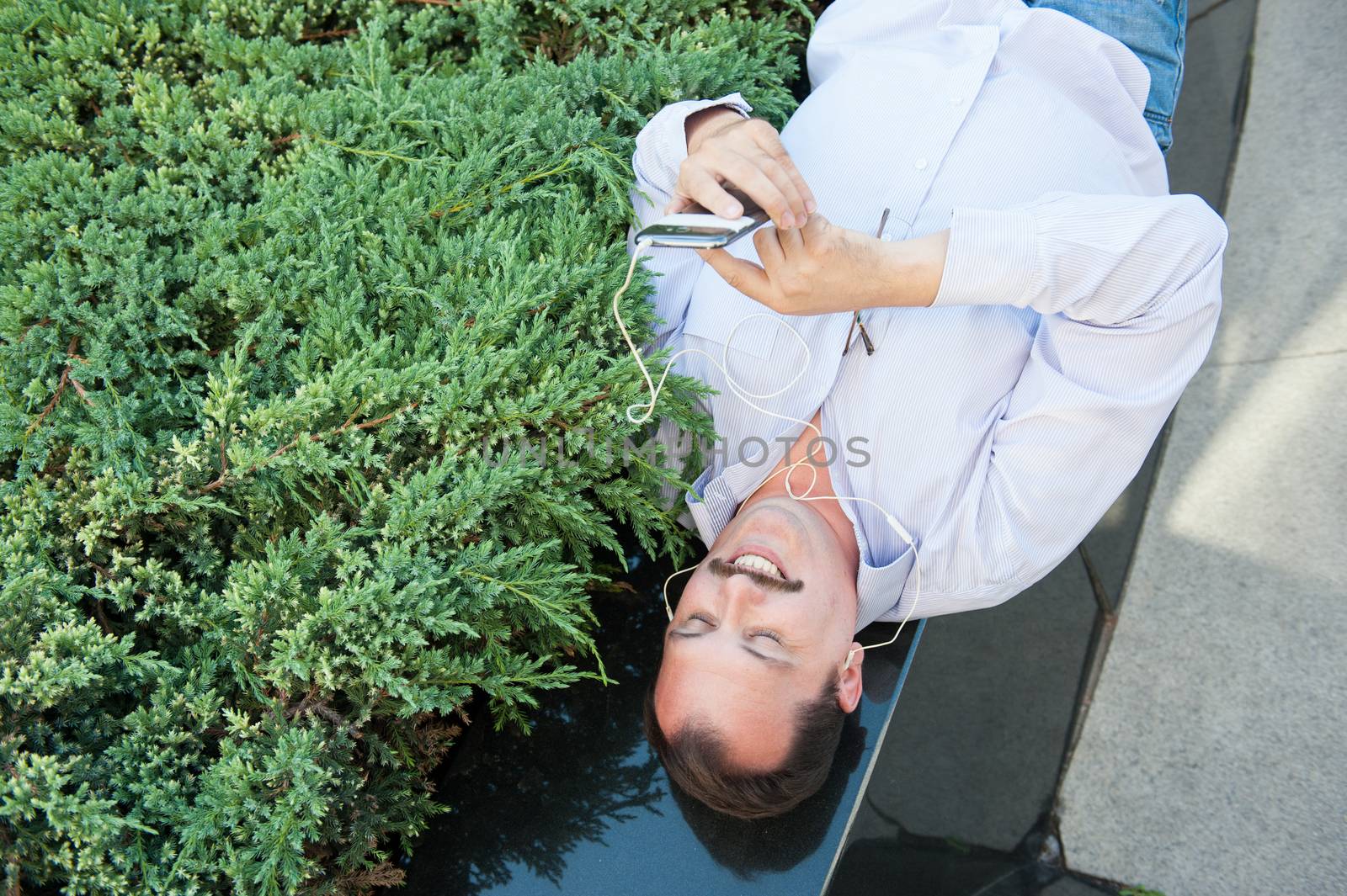 Urban man using smart phone outside using app on 4g wireless device wearing headphones. Lying on the Marble fence