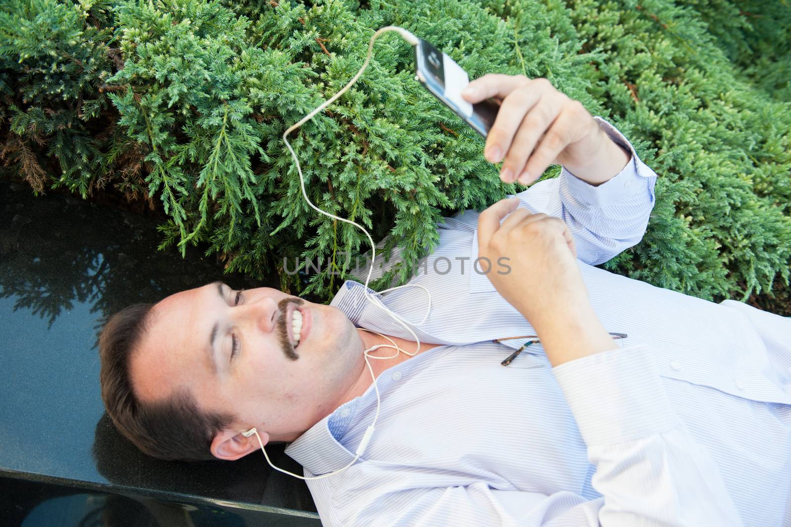 Urban man using smart phone outside using app on 4g wireless device wearing headphones. Lying on the Marble fence