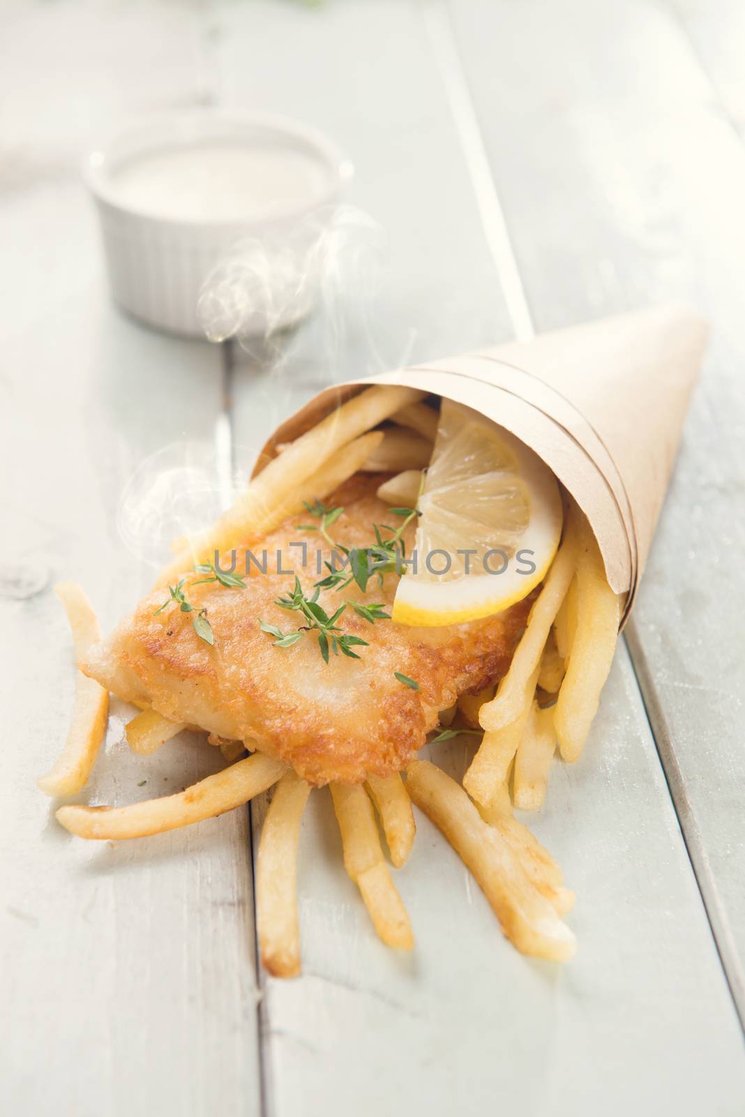 Fish and chips. Fried fish fillet with french fries wrapped by paper cone, on bright wooden background. Fresh cooked with hot smoke.