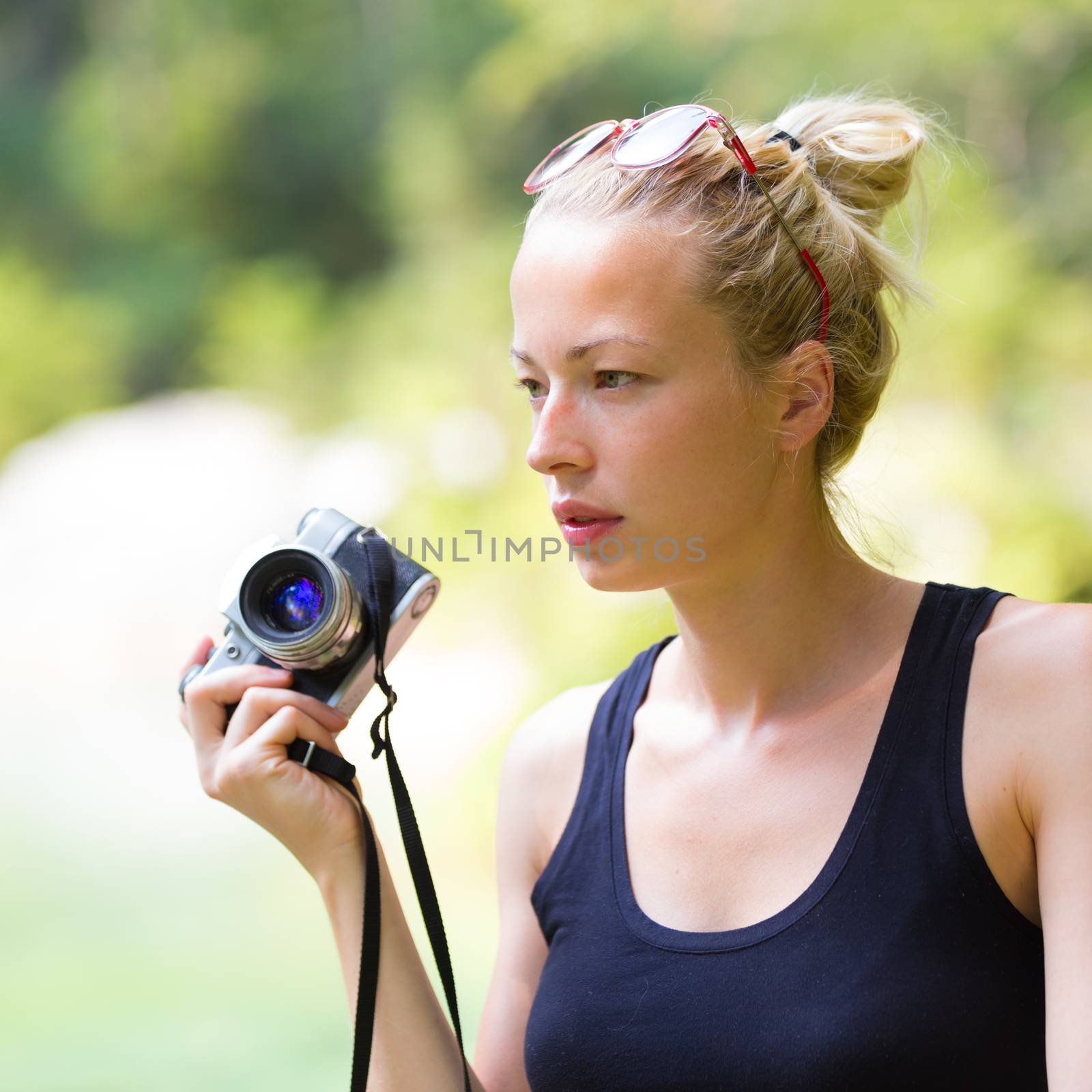 Portrait of eautiful blonde caucasian girl wearing sporty black sleeveless t-shirt, outdoors in nature, holding vintage camera in her hand. Square composition.
