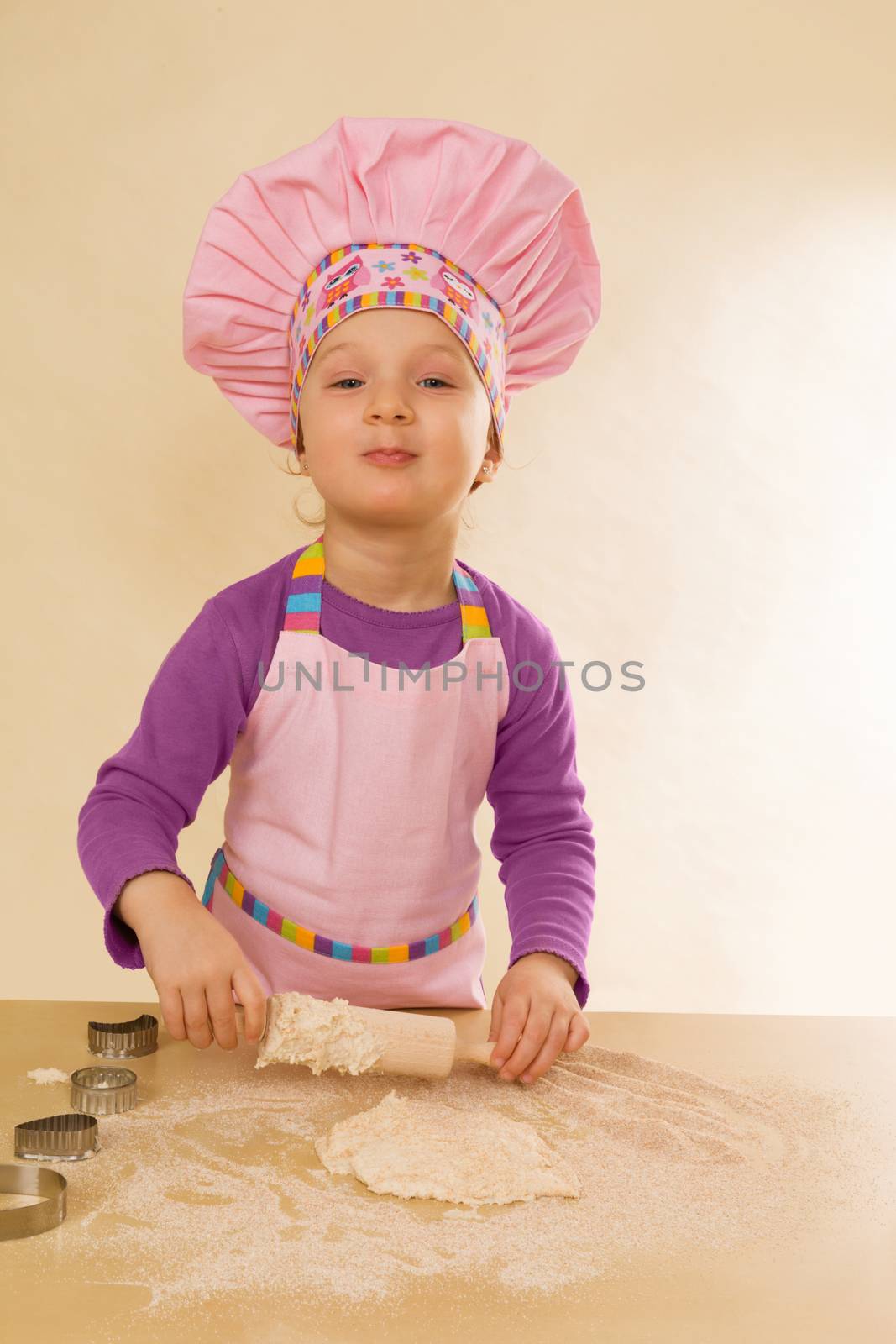 Little girl in chef hat and tablier with classic wooden rolling pin, dough and flour. Gastronomy and culinary cooking.