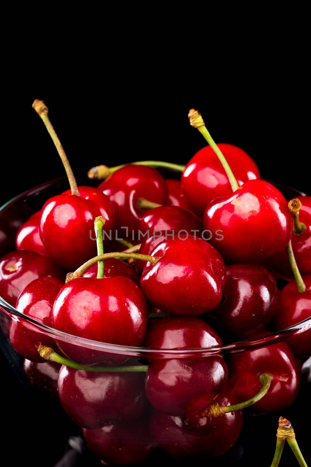 Juicy shiny cherries in a glass bowl on black background.