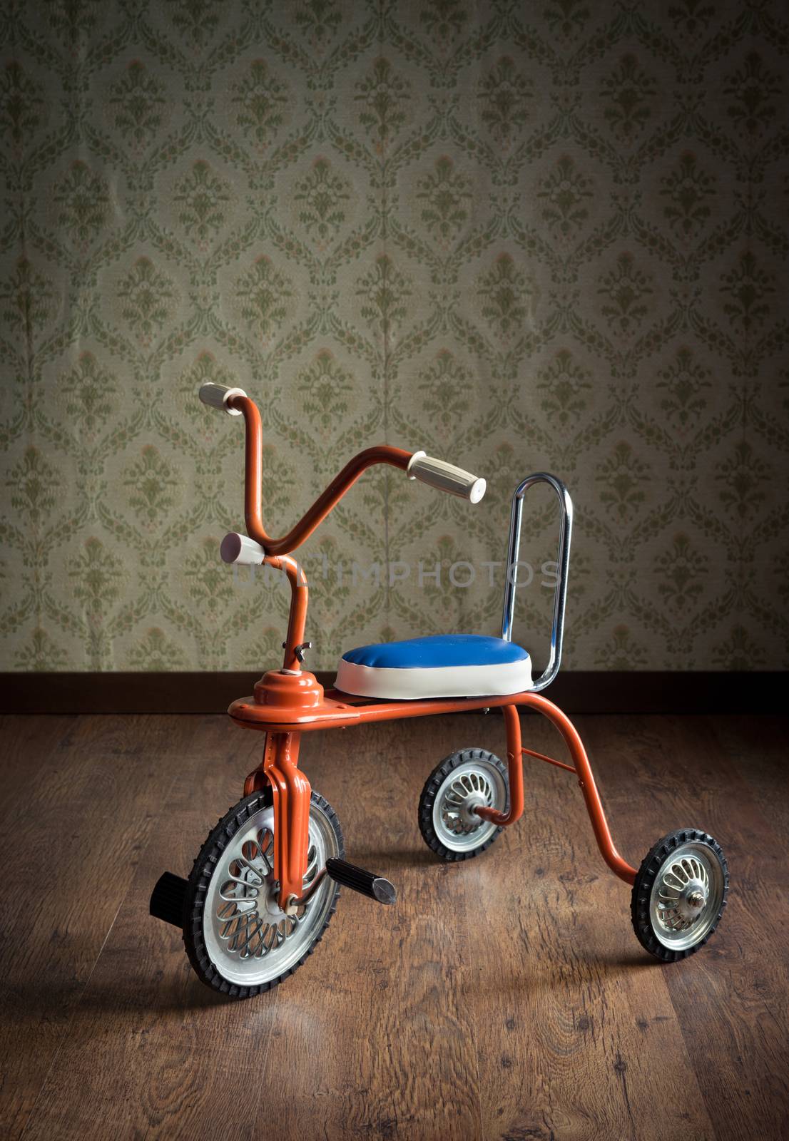Vintage orange tricycle on hardwood floor and vintage wallpaper on background.