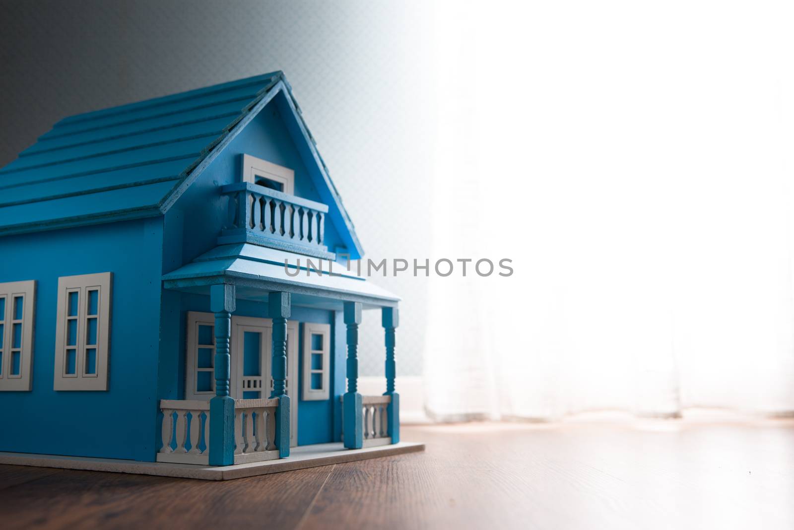 Blue wooden model house next to a window with curtain on wooden floor.