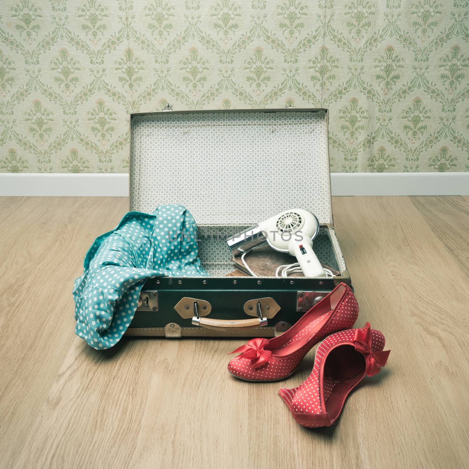 Open vintage suitcase with red shoes and dotted clothing, retro wallpaper on background.