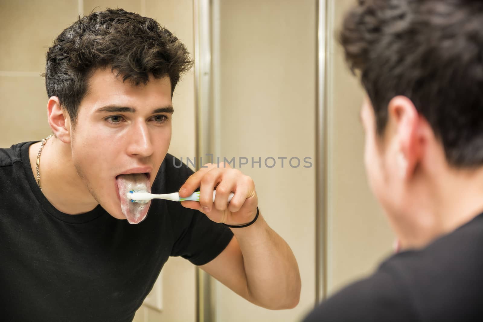 Headshot of attractive young man brushing teeth and cleaning tongue with toothbrush, looking at himself in mirror