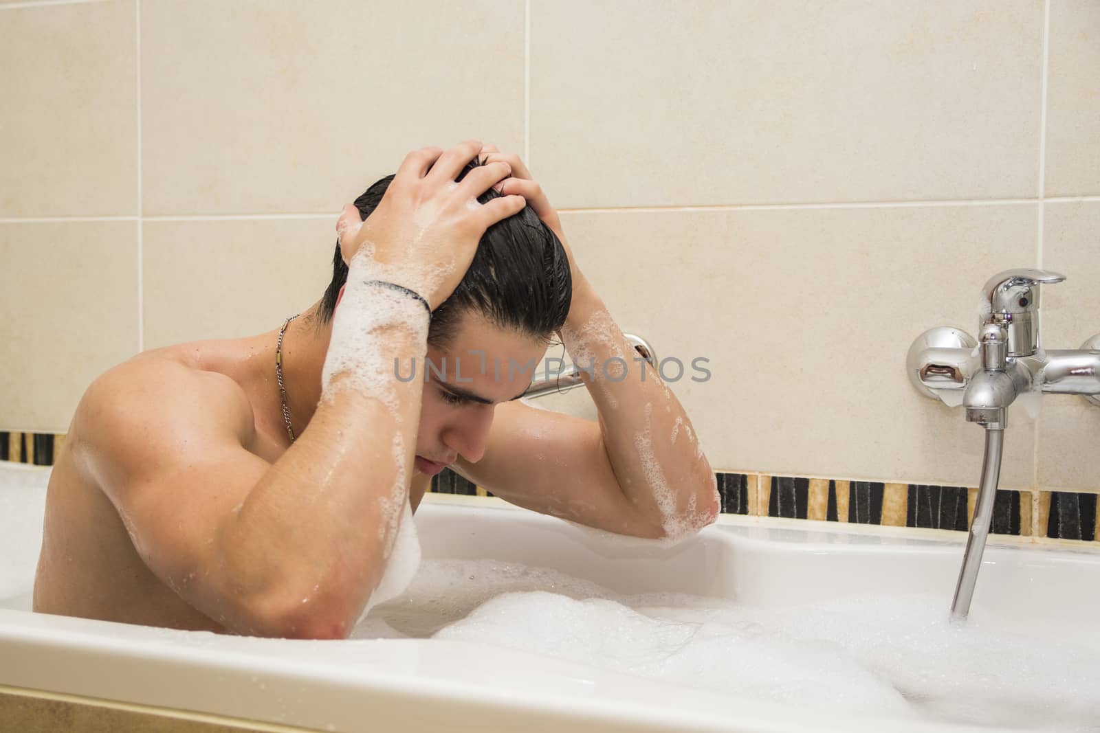 Handsome young man in bathtub at home having bath, washing body and hair with bathfoam and shampoo