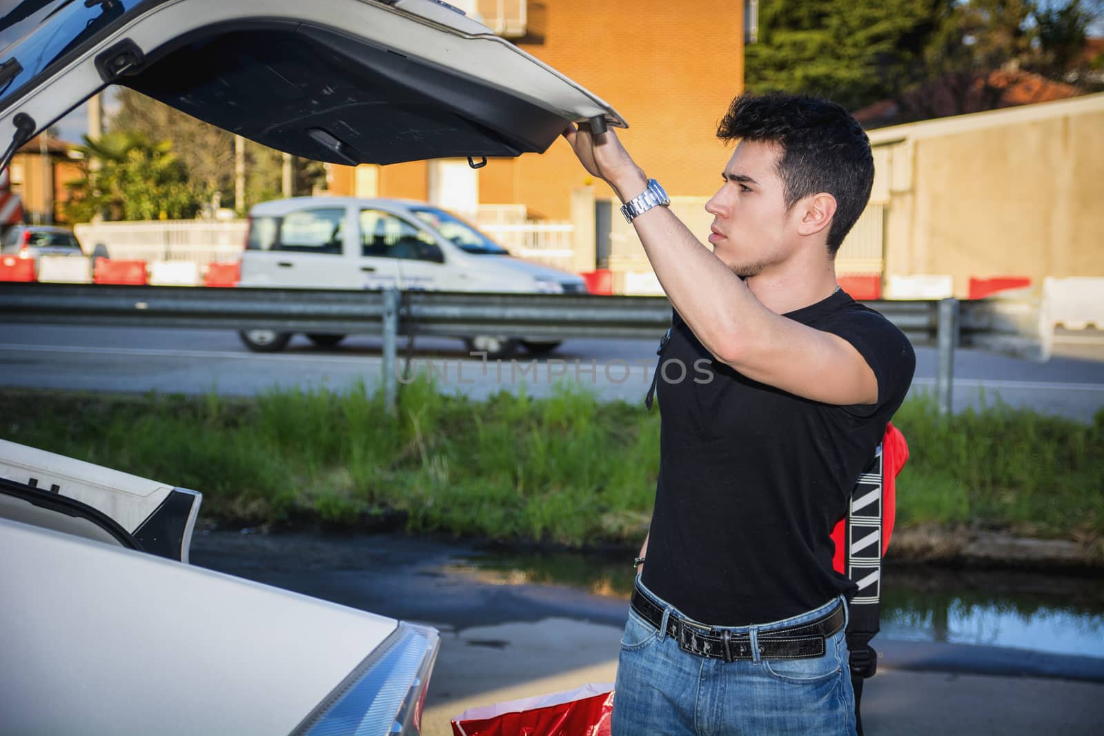 Young man taking luggage and bag out of car trunk by artofphoto