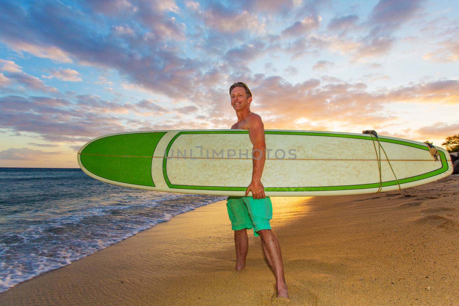 Athletic upbeat adult holding his surfboard on beach at sunset