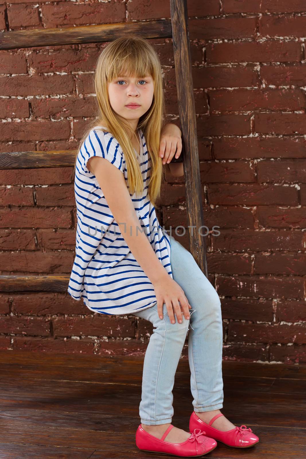 Girl 6 years old in jeans and a vest sits on a ladder near a brick wall