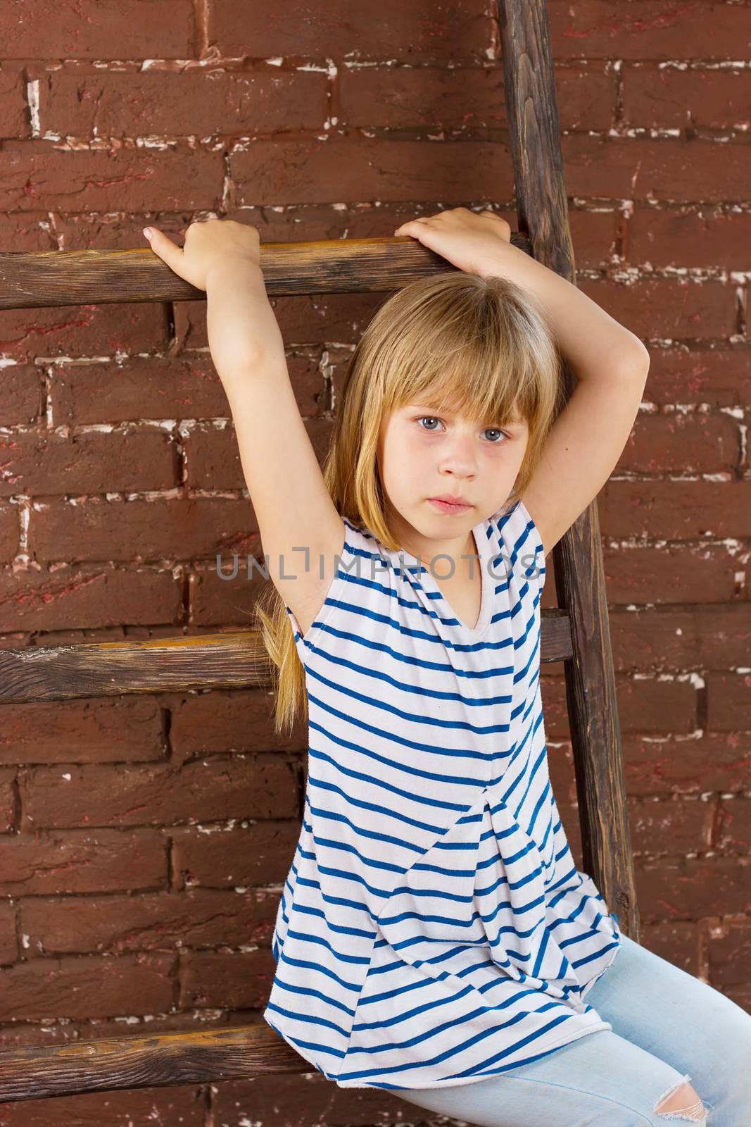 Girl 6 years old in jeans and a vest sits on a ladder near a brick wall. Vertical framing