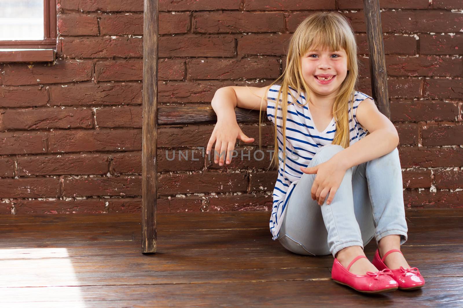 Girl 6 years old in jeans and a vest sits on the floor next to a brick wall and filled with laughter