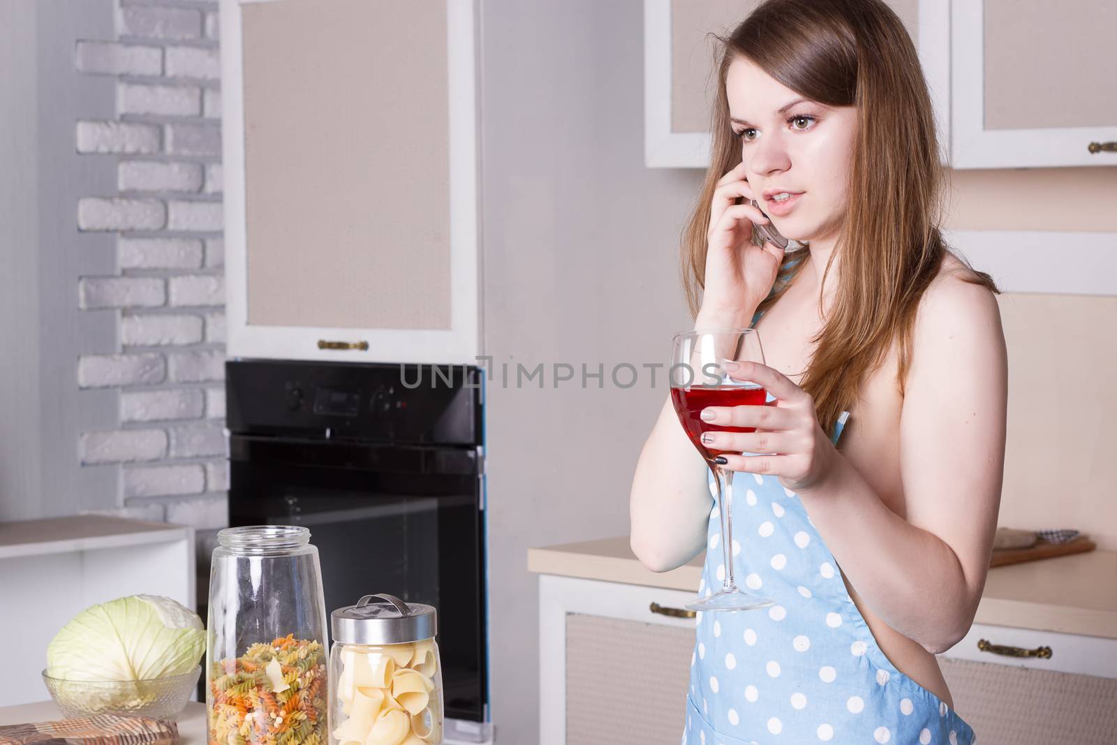 girl in the kitchen wearing an apron over his naked body with a glass of wine on the phone