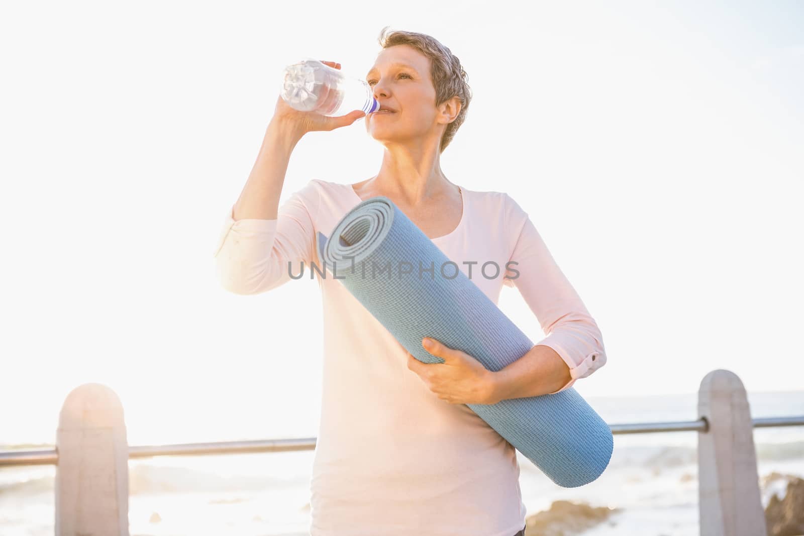 Sporty woman with exercise mat drinking water at promenade