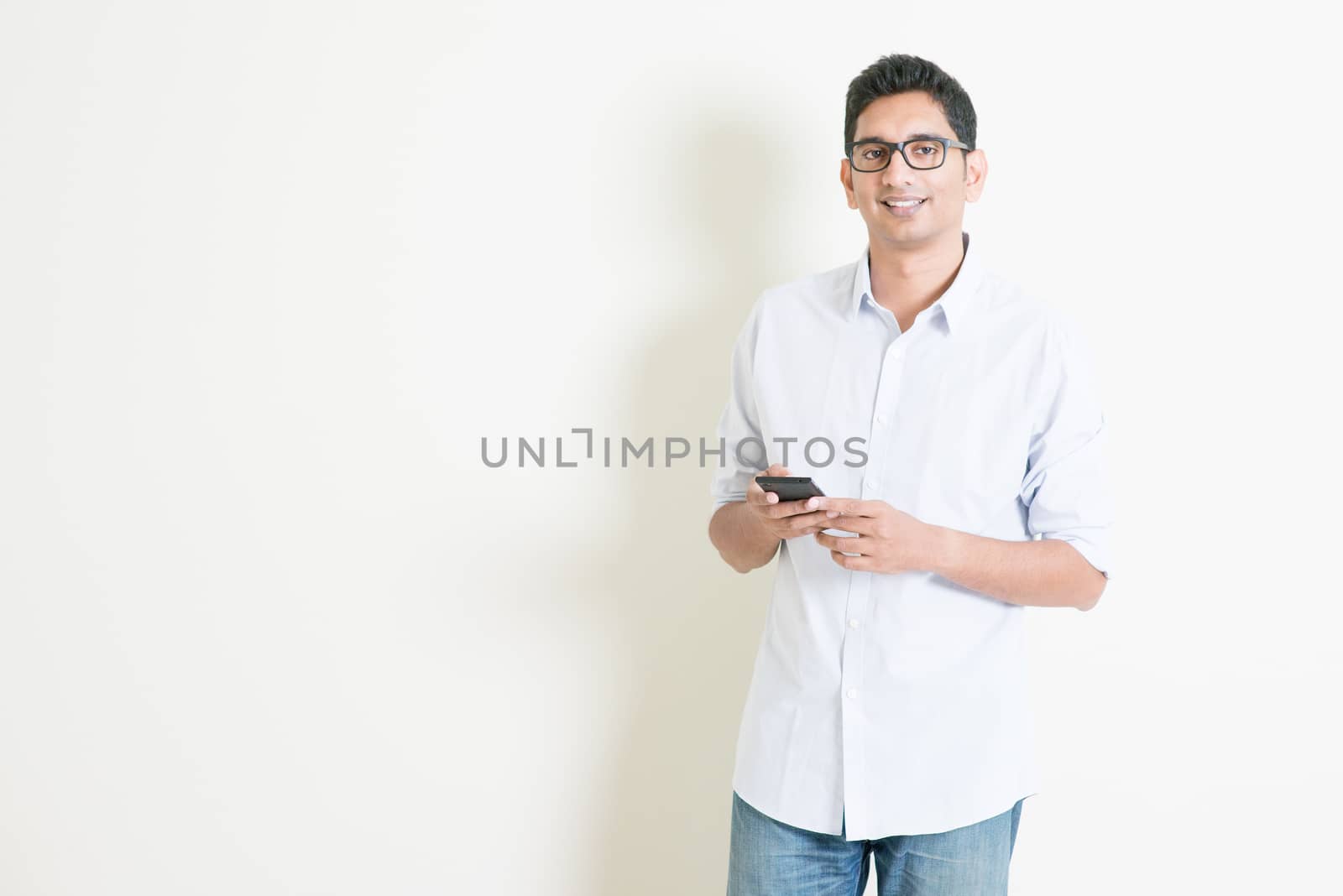 Portrait of handsome casual business Indian man using smartphone, social media concept, standing on plain background with shadow, copy space at side.