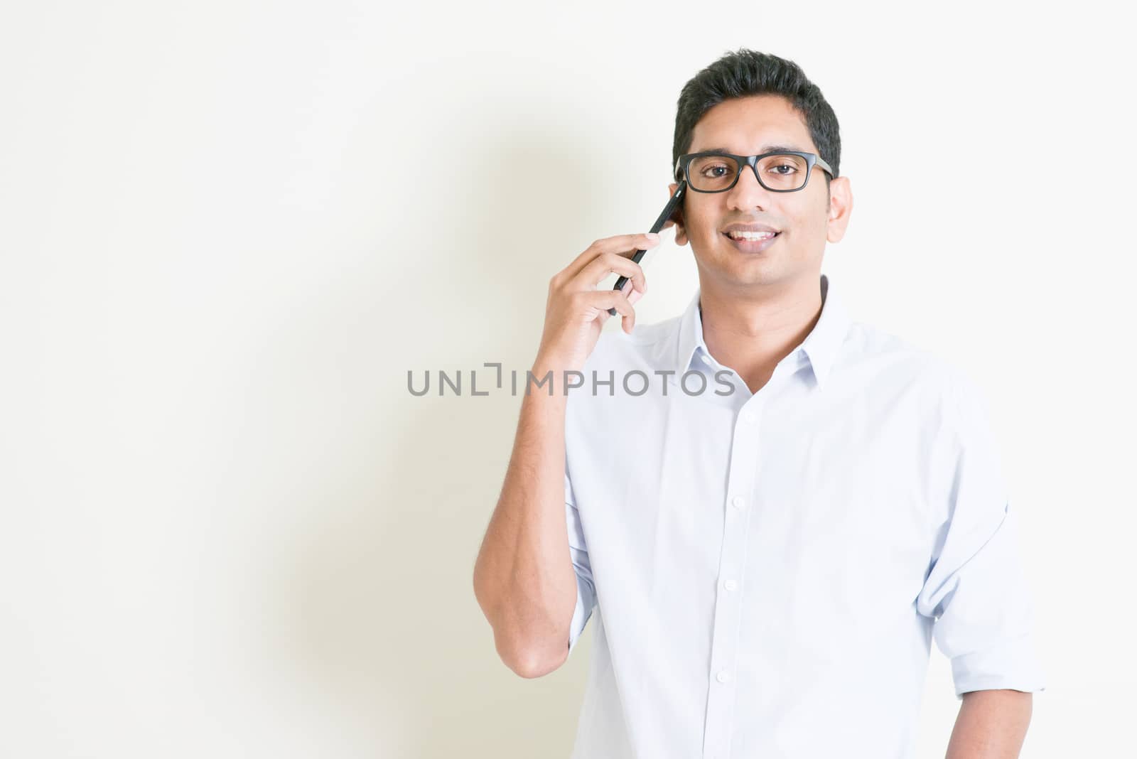 Portrait of handsome casual business Indian man talking on smartphone, standing on plain background with shadow, copy space at side.