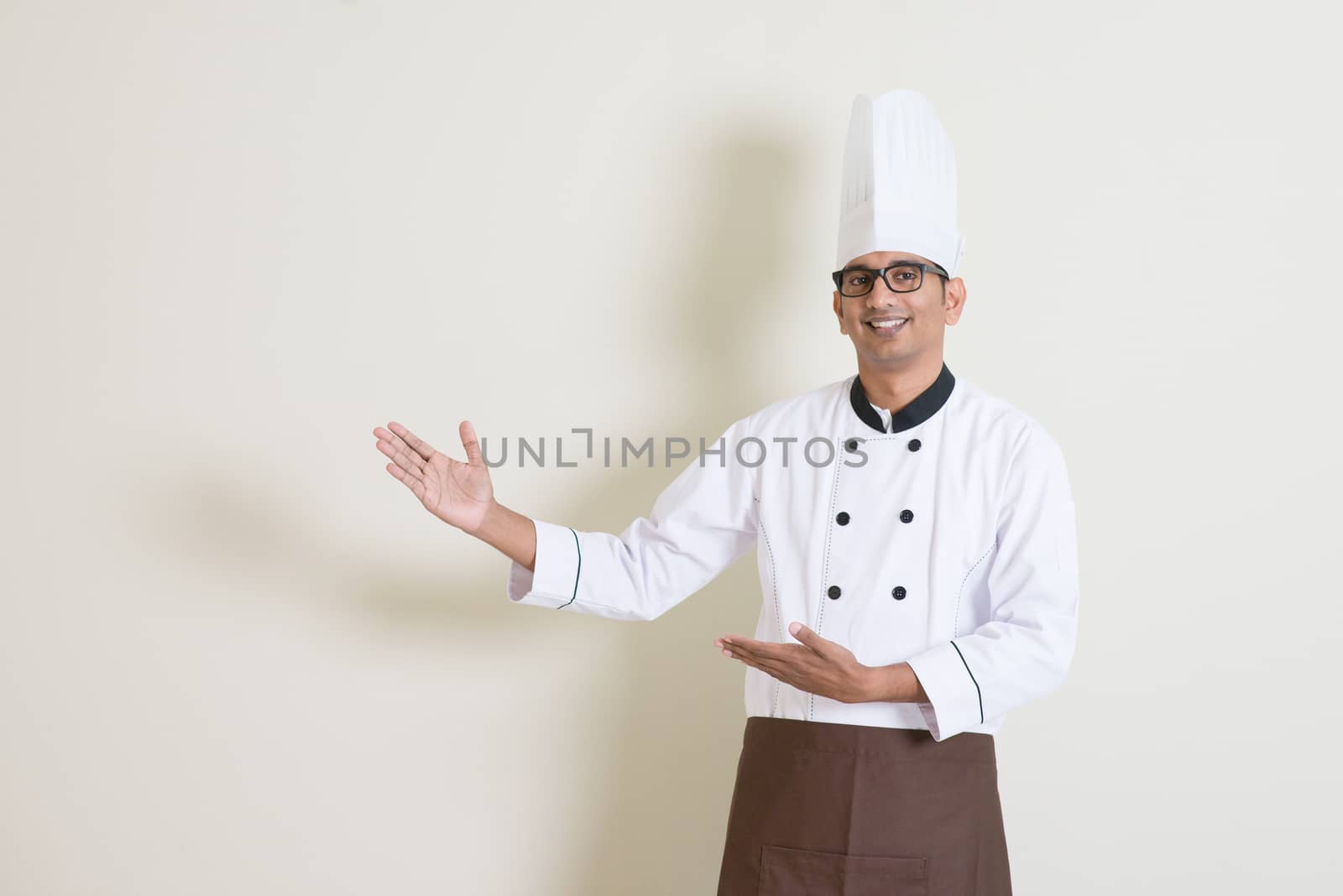 Portrait of handsome Indian male chef in uniform hands gesturing showing something and smiling, standing on plain background with shadow, copy space at side.