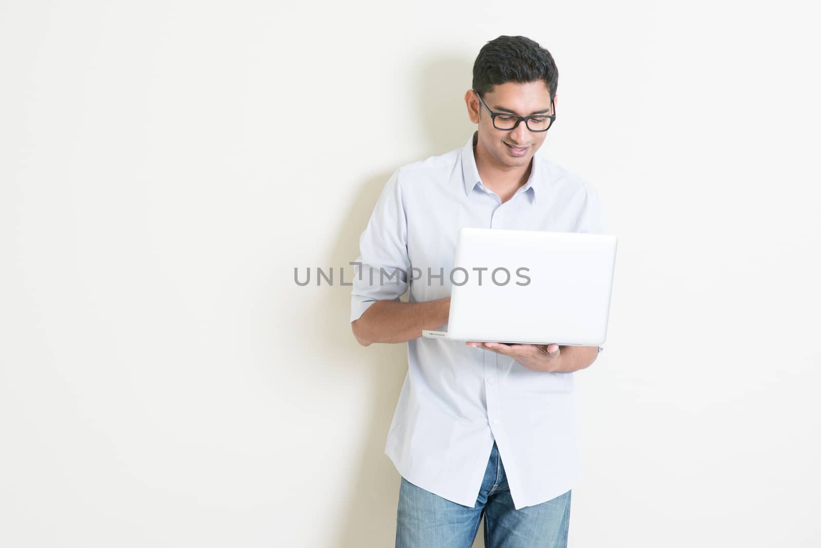 Portrait of handsome casual business Indian man using laptop computer, standing on plain background with shadow, copy space at side.