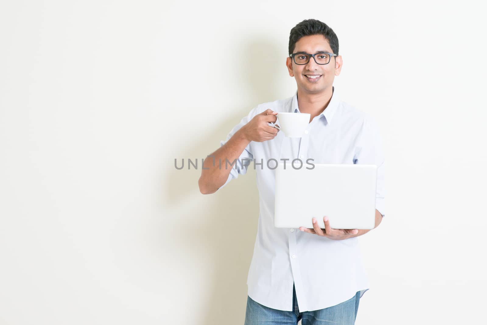 Portrait of handsome casual business Indian guy drinking coffee while using laptop computer, standing on plain background with shadow, copy space at side.