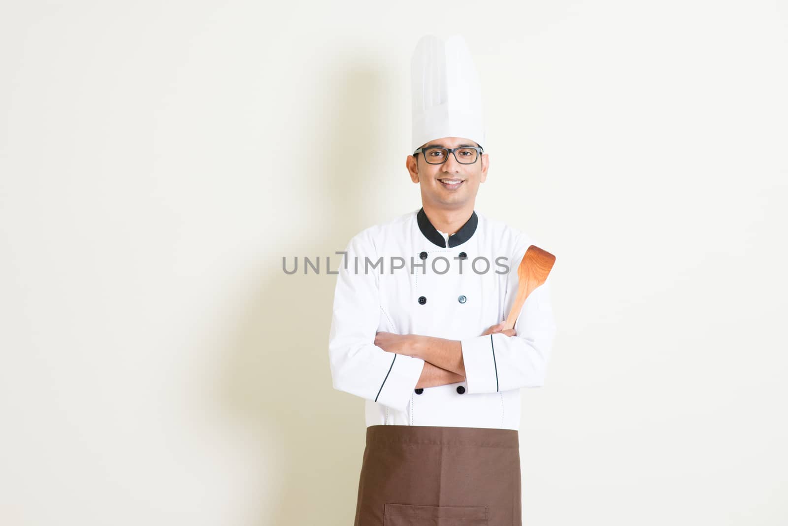 Portrait of handsome Indian male chef in uniform hand holding spatula and smiling, standing on plain background with shadow, copy space on side.
