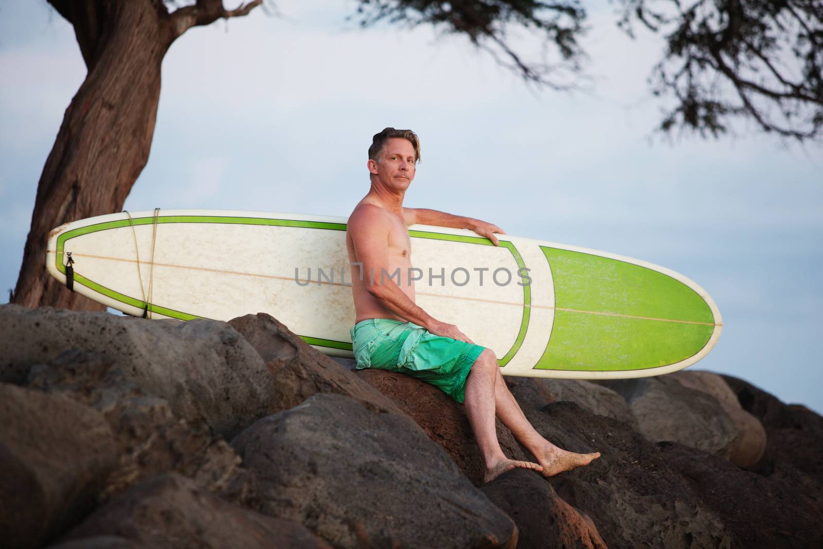 Single adult surfer male outdoors sitting with surfboard on rocks