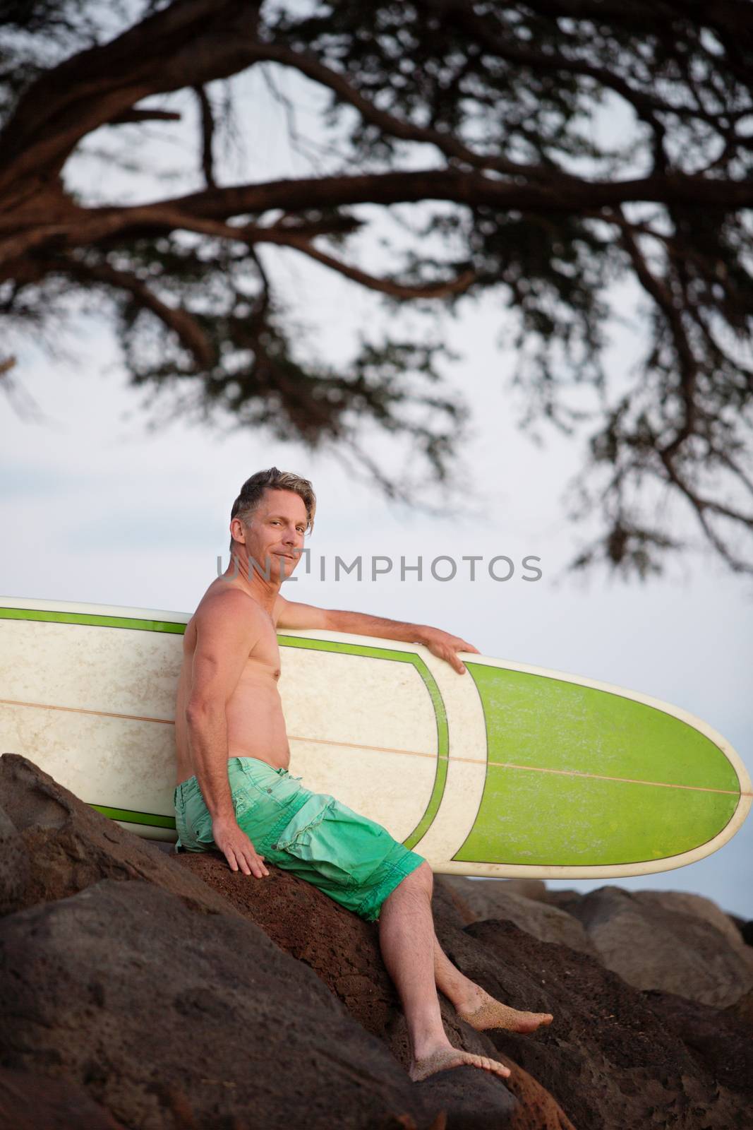 Single man sitting with blank surfboard on rocks outdoors