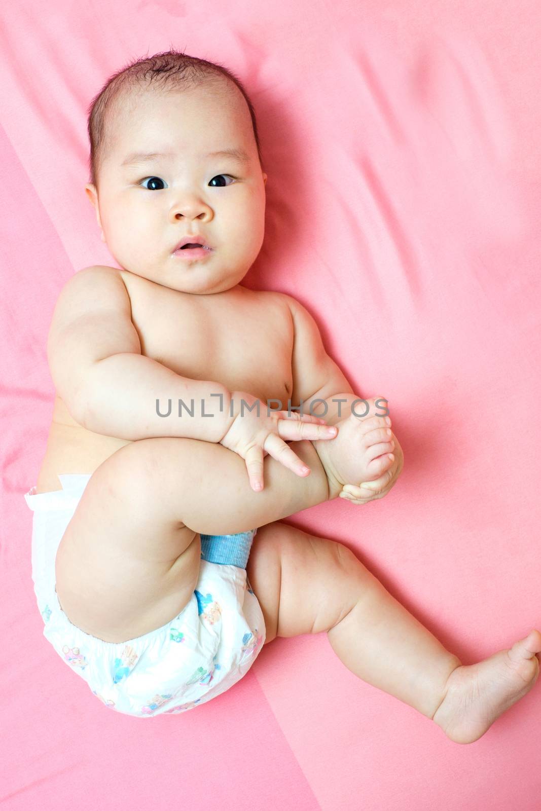 Asian baby girl with diaper touching her foot on pink bed and she looking at the camera
