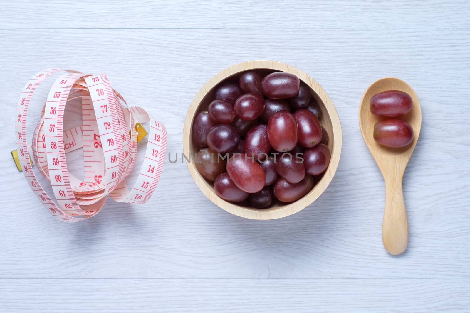 red grapes in wooden bowl and spoon and tape measure next to them. they on white wooden table. Clear light. This photo use for descript weight loss, diet, eat clean