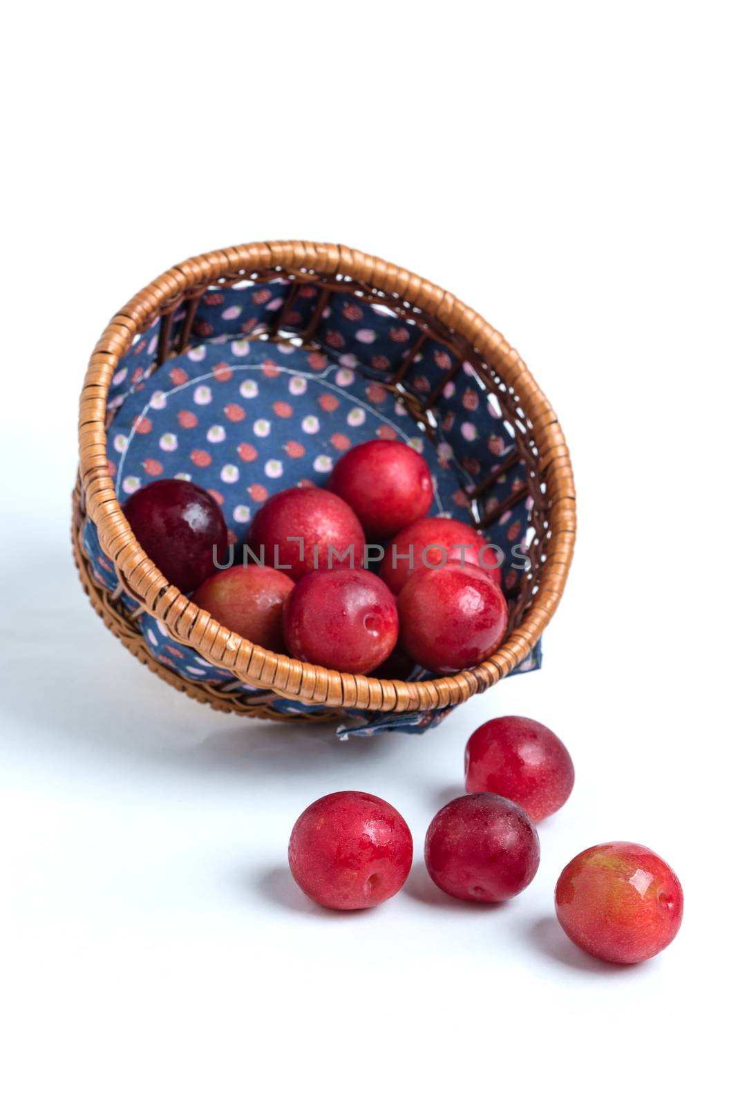 Red plums pouring out of a wicker basket with a blue cloth onto a white background.