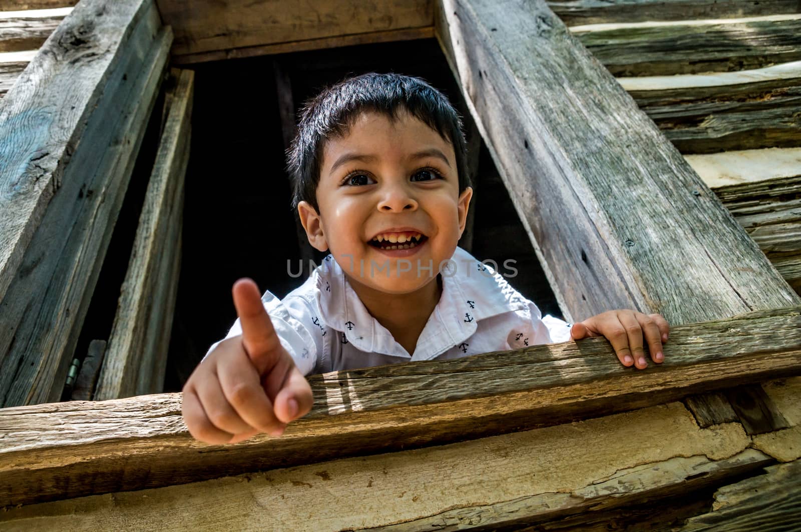 Latino child looking out the window of a wooden house or shed, pointing at the camera and smiling