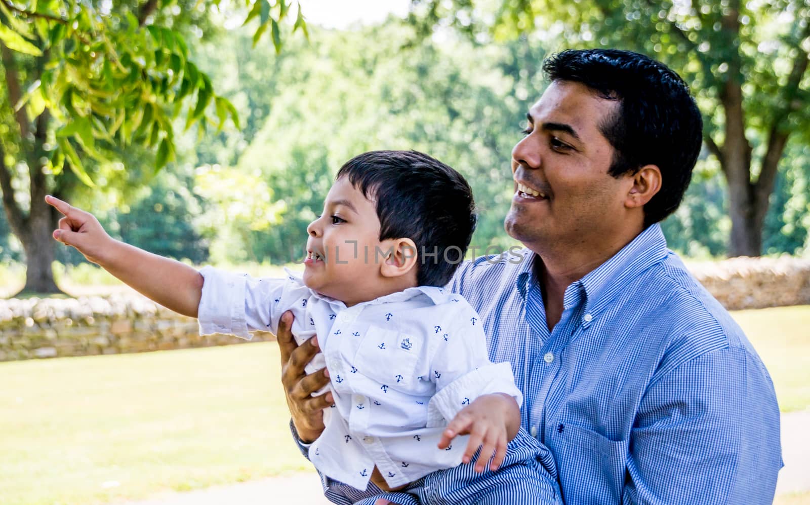 Latino father holding his son outside in a park with trees in the background. The boy is pointing at something in the distance and the father is smiling.