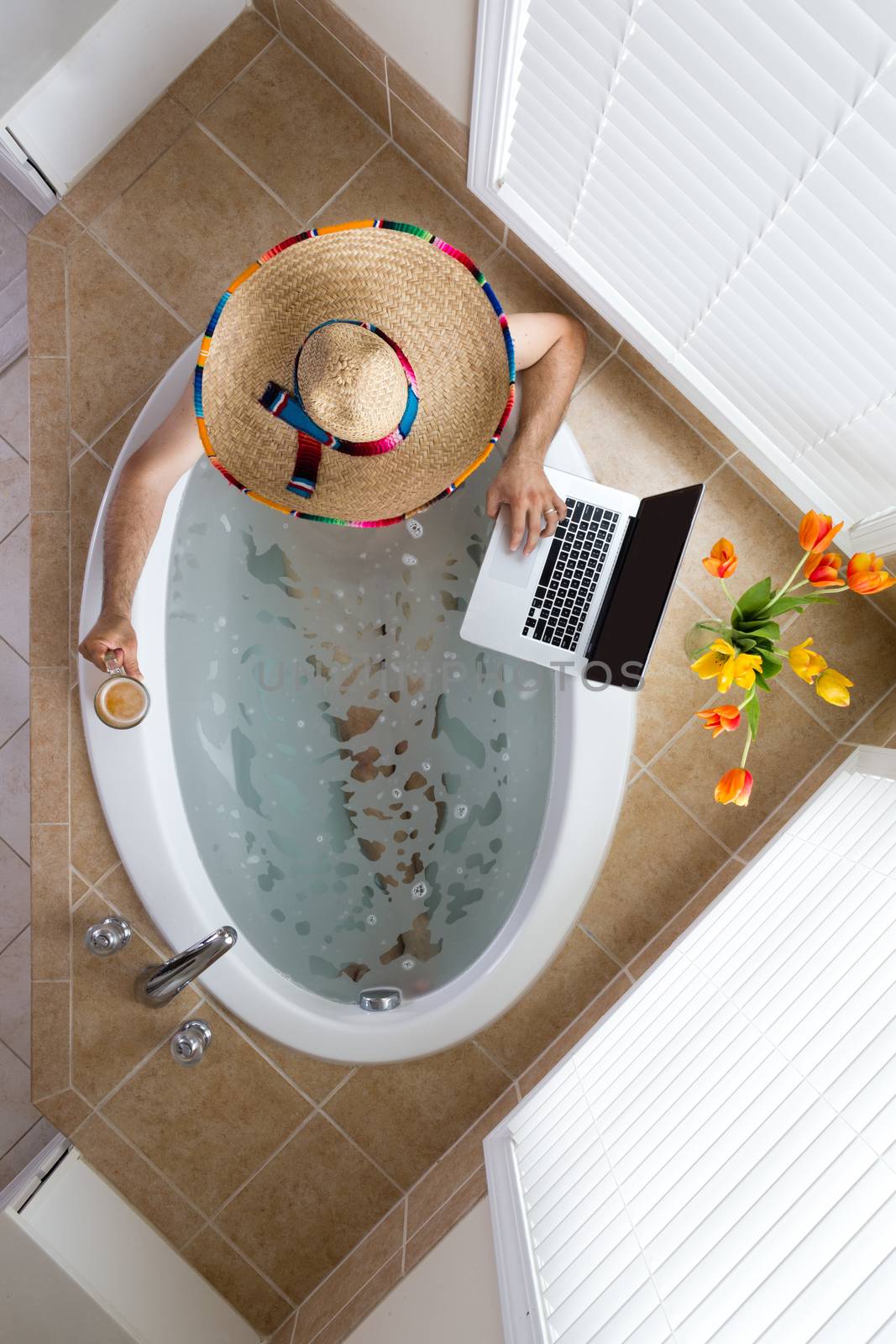 Man relaxing in a bath in a Mexican sombrero with a beer in one hand and using his laptop computer with the other, conceptual of a tourist on vacation in Mexico, overhead view
