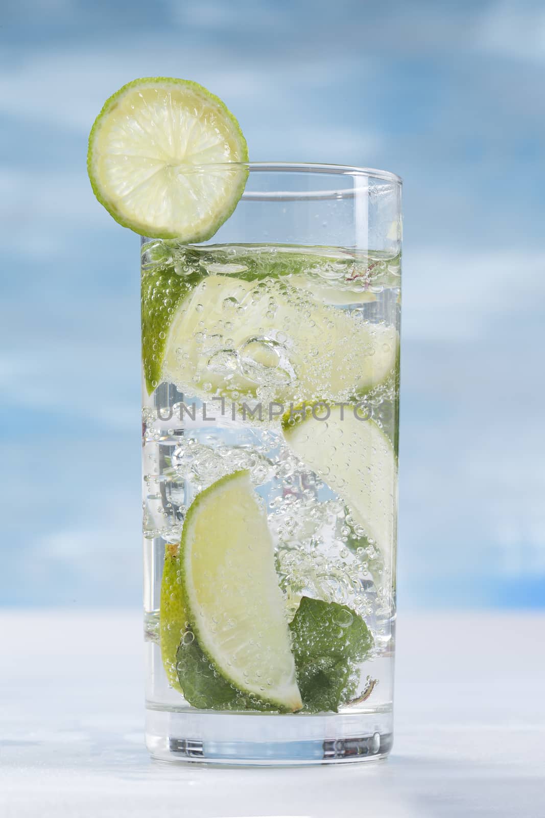 Glass of sparkling water with ice cubes garnished with a slice of lime and mint on blue sky background