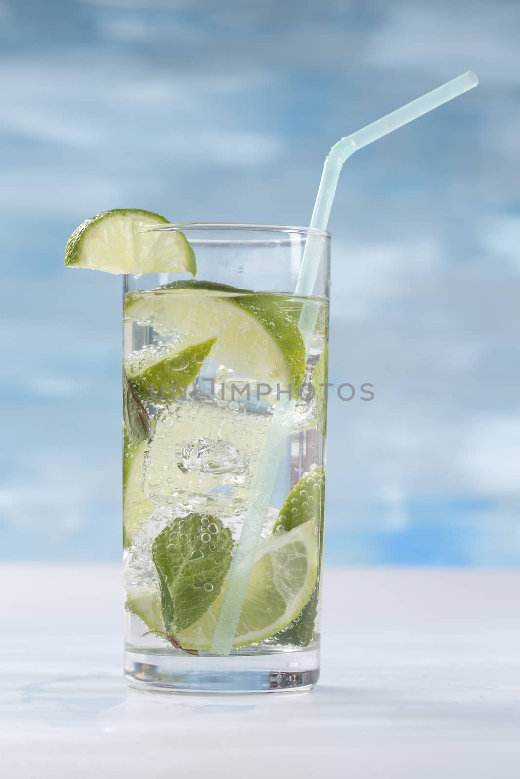 Glass of sparkling water with ice cubes garnished with a slice of lime and mint on blue sky background