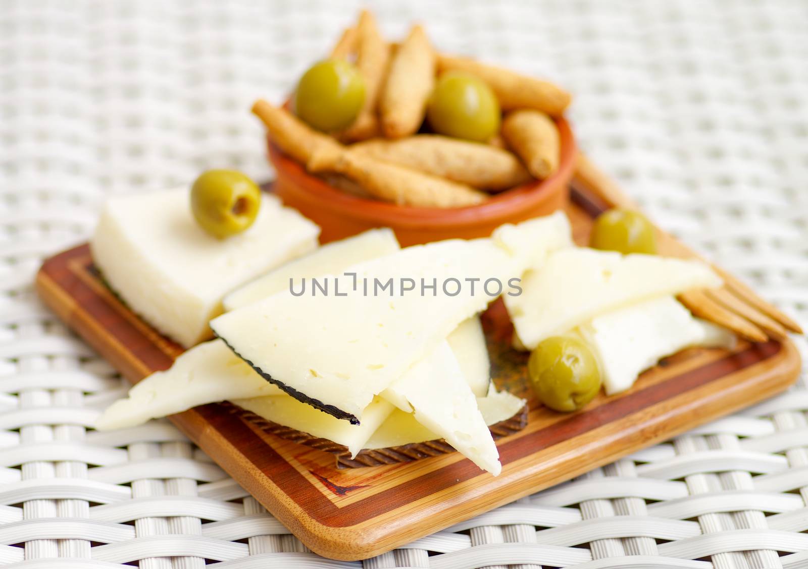 Various Goat Cheeses with Bread Sticks and Green Olives on Wooden Plate closeup on Wicker background. Focus on Foreground