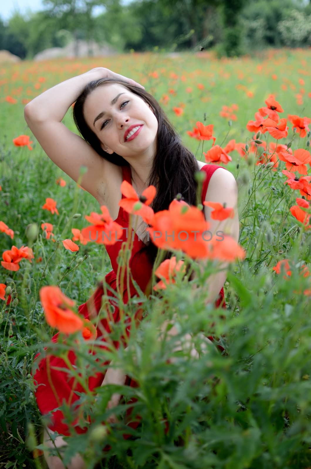 Beautiful polish girl surrounded by field full of poppies. Young female model with charming smile, wearing red dress.