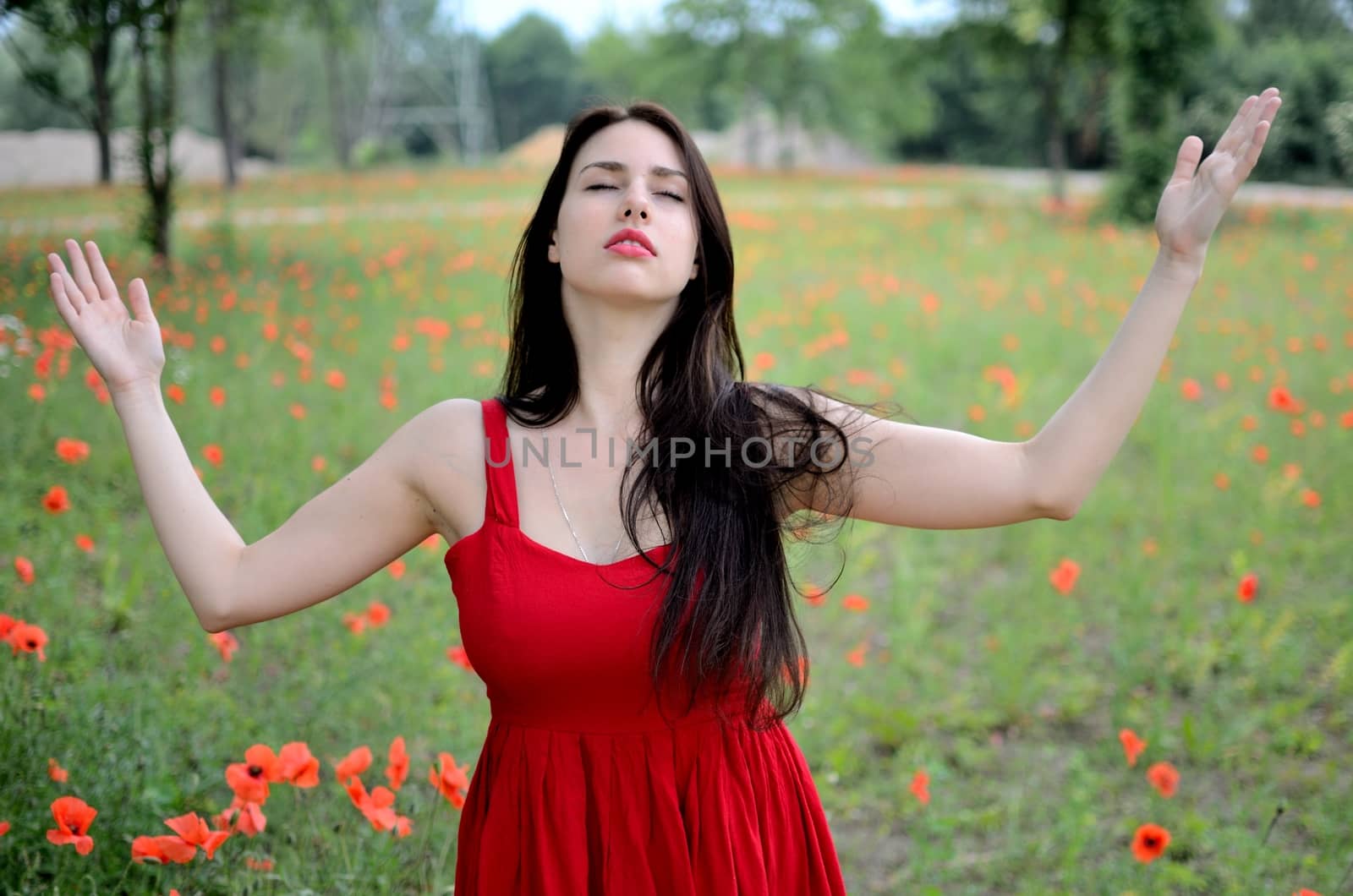 Female model with red dress. Girl meditating in open field, meadow with poppies in background. Model with closed eyes and open arms.