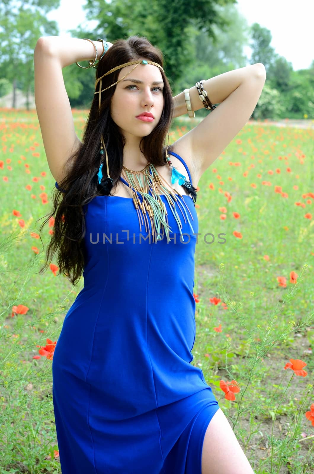 Brunette model wearing long blue dress with simple jewelry. Girl stands in meadow surrounded by poppies.