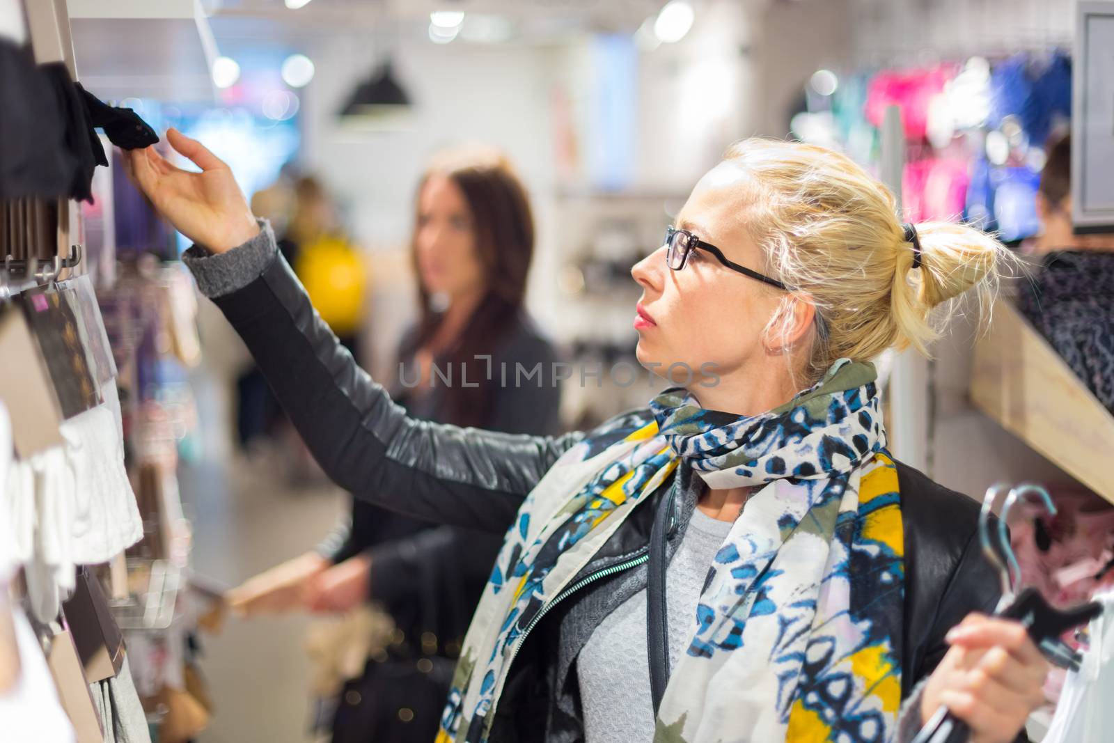 Woman shopping clothes. Shopper looking at clothing indoors in store. Beautiful blonde caucasian female model wearing black glasses, casual black leather jacket an colorful scarf.