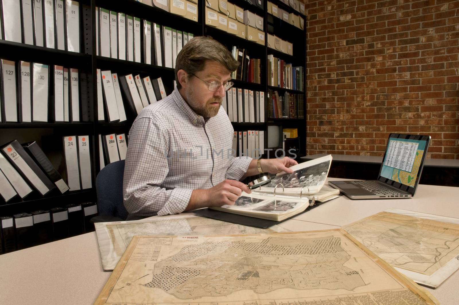 Researcher in archive, searching through maps and photographs. [Maps and photographs shown are public domain. Image on computer was created for this photograph. Model & Property Released.]